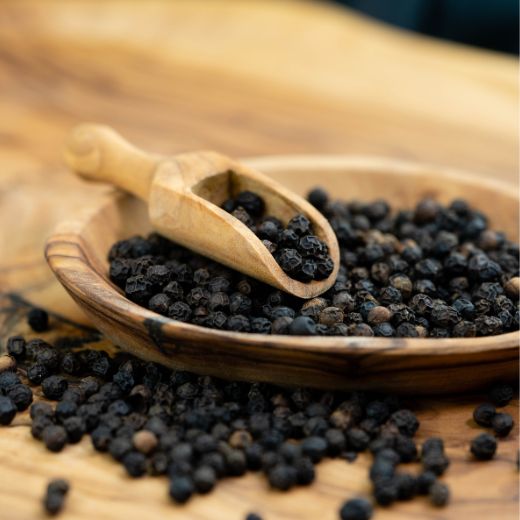 Whole black peppercorns arranged in a wooden scoop and bowl on a warm wooden surface.