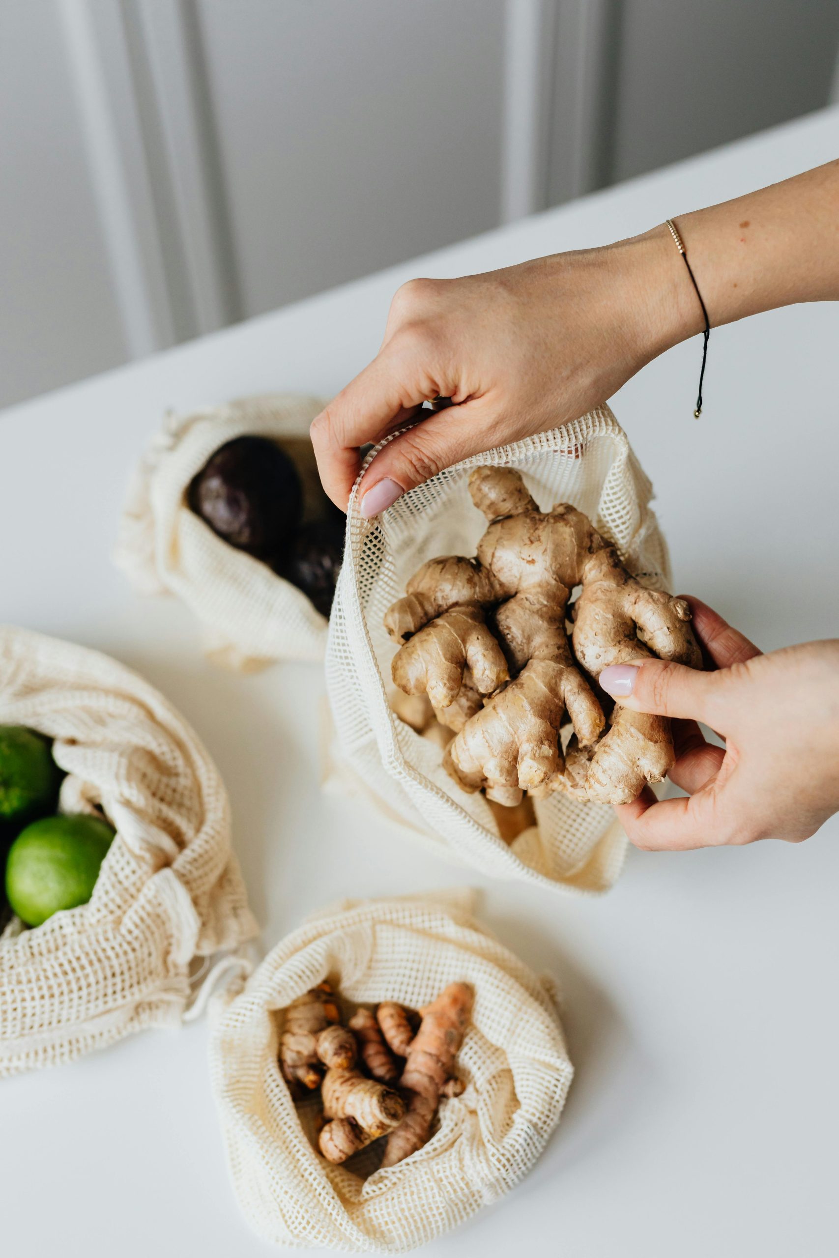 Fresh Ginger in Reusable Produce Bag Hands placing fresh ginger root into a reusable mesh produce bag, with turmeric and limes nearby on a clean white surface.