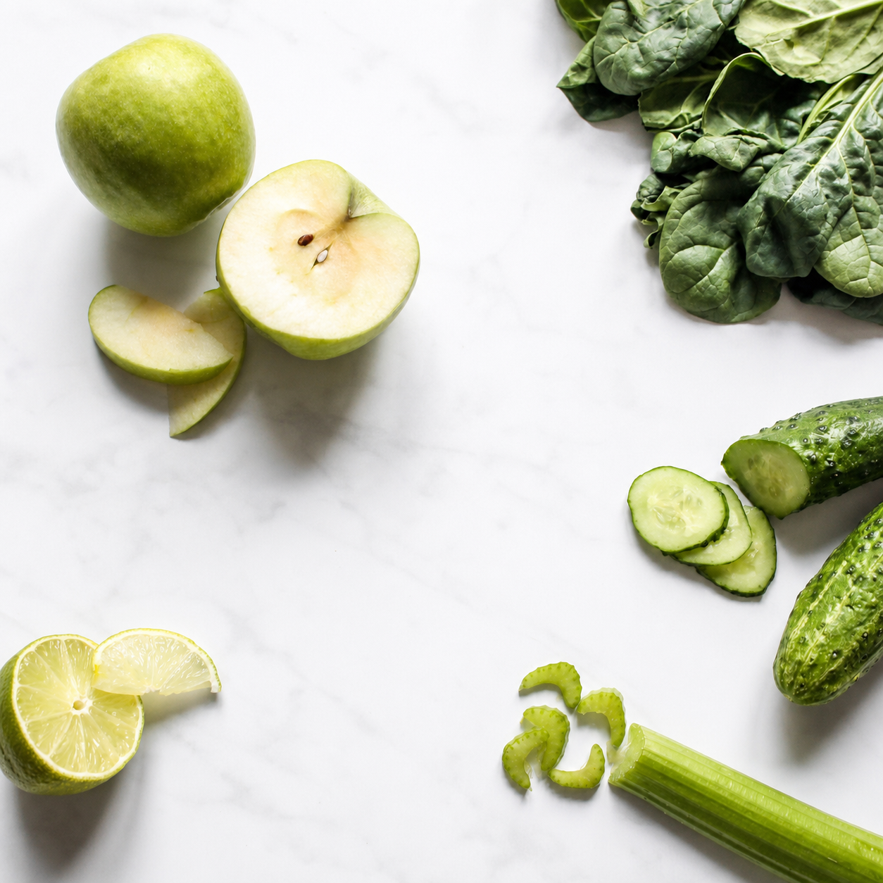Green apples, spinach, cucumbers, lime and celery arranged around an empty white marble surface.